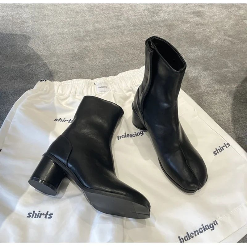 par de botas pretas sobre uma sacola de tecido branco — vista de cima em ângulo, duas botas pretas com bico split toe bem visível, salto grosso redondo, cabedal liso, apoiadas sobre fundo de tecido cinza. Contexto lifestyle/unboxing sofisticado.