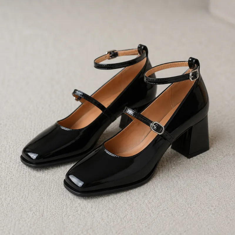 par de sapatos sobre tapete bege claro. Bico quadrado com cantos suavizados em verniz preto brilhante, duas tiras com fivelas prateadas (tira Mary Jane no peito do pé + tira no calcanhar), interior bege caramelo bem visível, salto grosso quadrado preto. Foto editorial limpa que mostra o produto completo com muito detalhe.