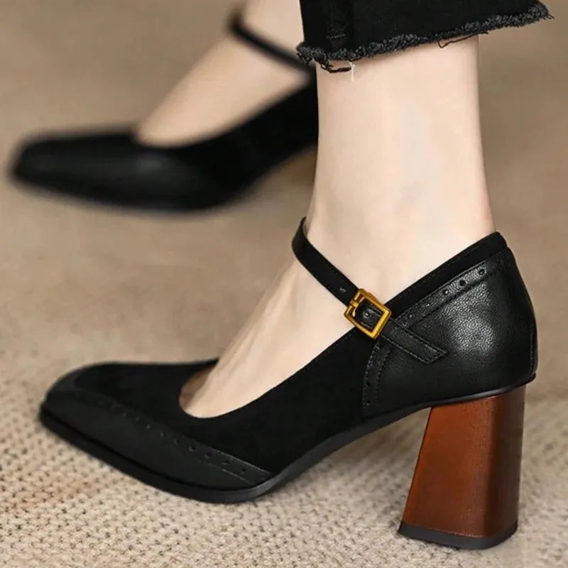 Close-up nos pés em uso real — vista frontal/lateral do scarpin preto no pé, calça preta no topo, tira com fivela dourada bem visível, detalhes brogue perfurados no bico quadrado, salto grosso flare em marrom, sobre tapete de fibra natural. Look total black elegante.